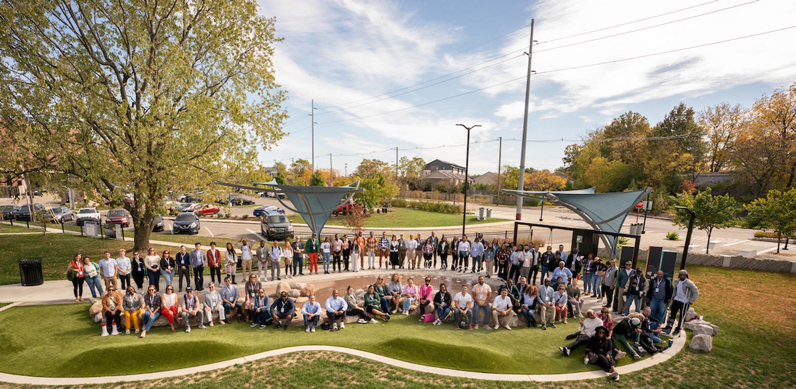 A large group of people gathers in an outdoor park, some sitting on grass while others stand. The day is sunny, with trees and cars visible in the background.