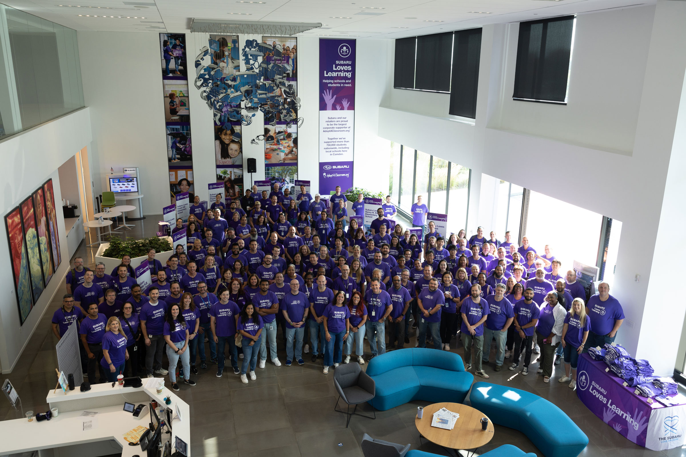 A large, diverse group of people wearing matching purple "Subaru Loves Learning" t-shirts, smiling in a lobby.