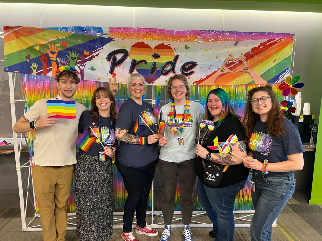 A group of six people holding rainbow flags and smiling under a vibrant Pride banner and rainbow decor.