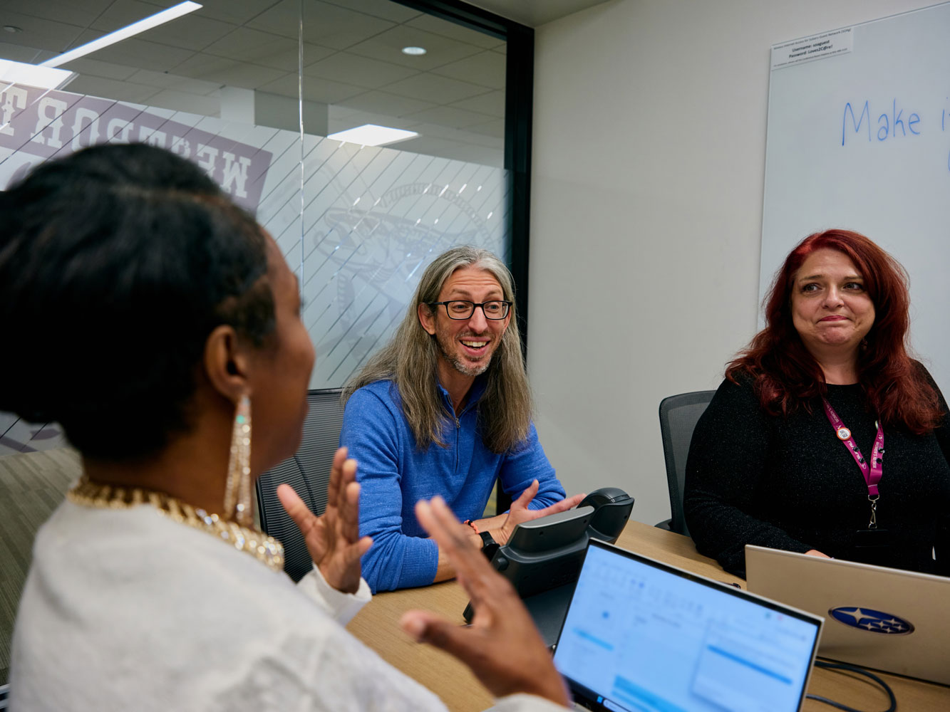 Three people in a meeting room with laptops, engaged in a lively discussion. One smiles warmly, while another gestures expressively.