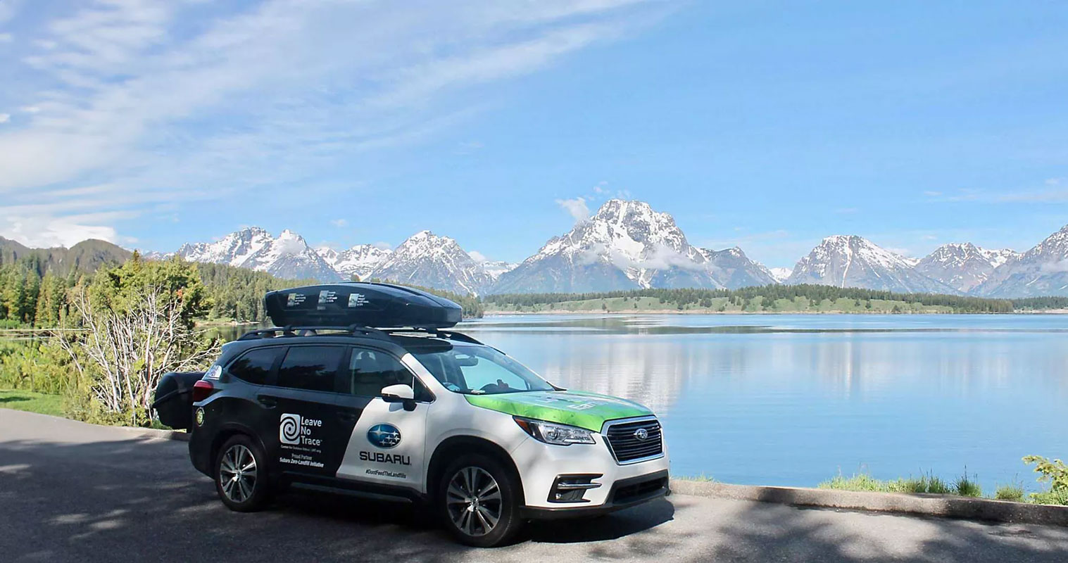 A Subaru SUV parked by a serene lake with snow-capped mountains in the background. The car displays "Leave No Trace" and environmental graphics.