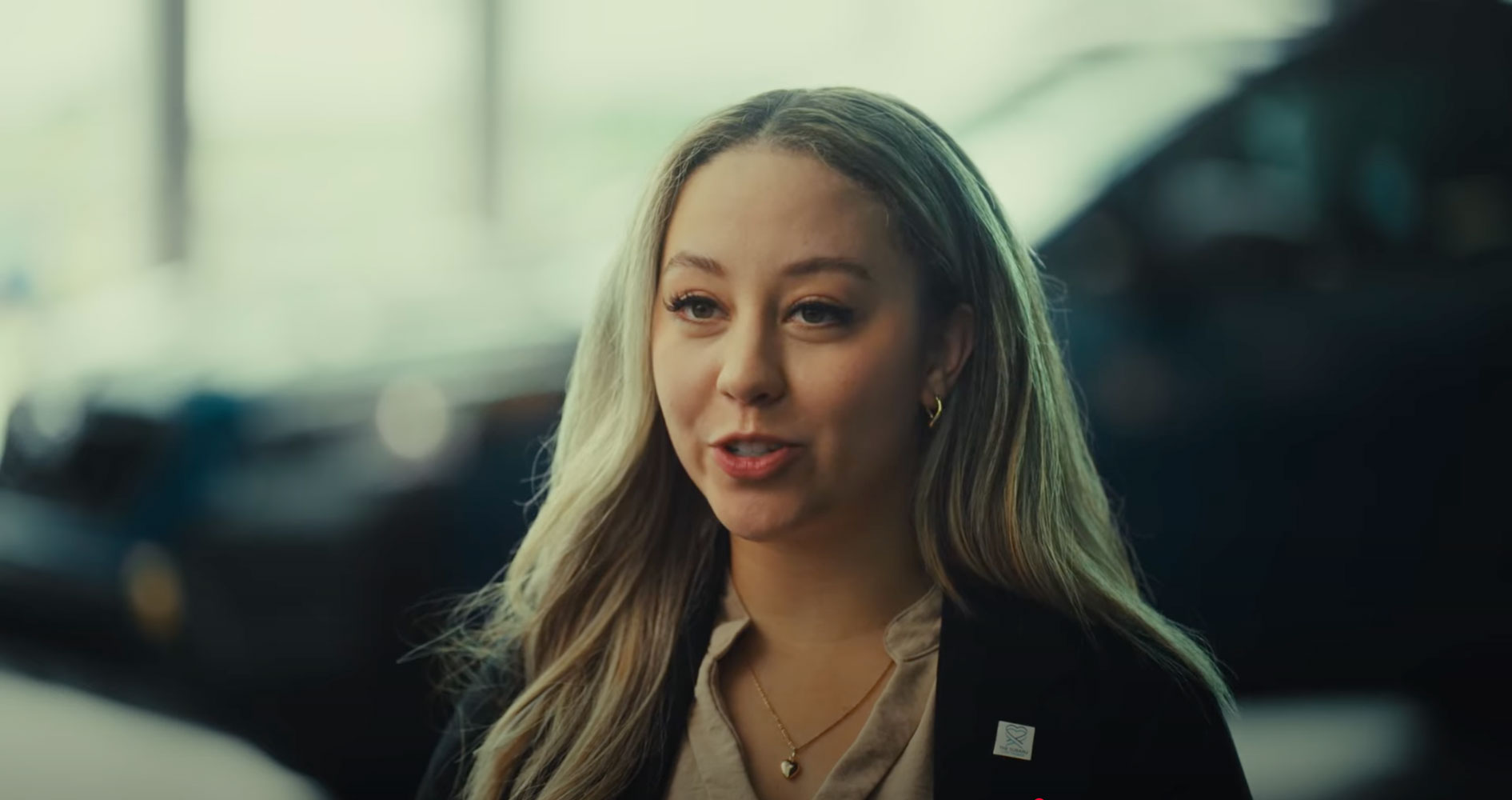 A woman with long blonde hair smiles warmly in front of a softly blurred background. She wears a tan blouse, black blazer, and gold heart necklace.