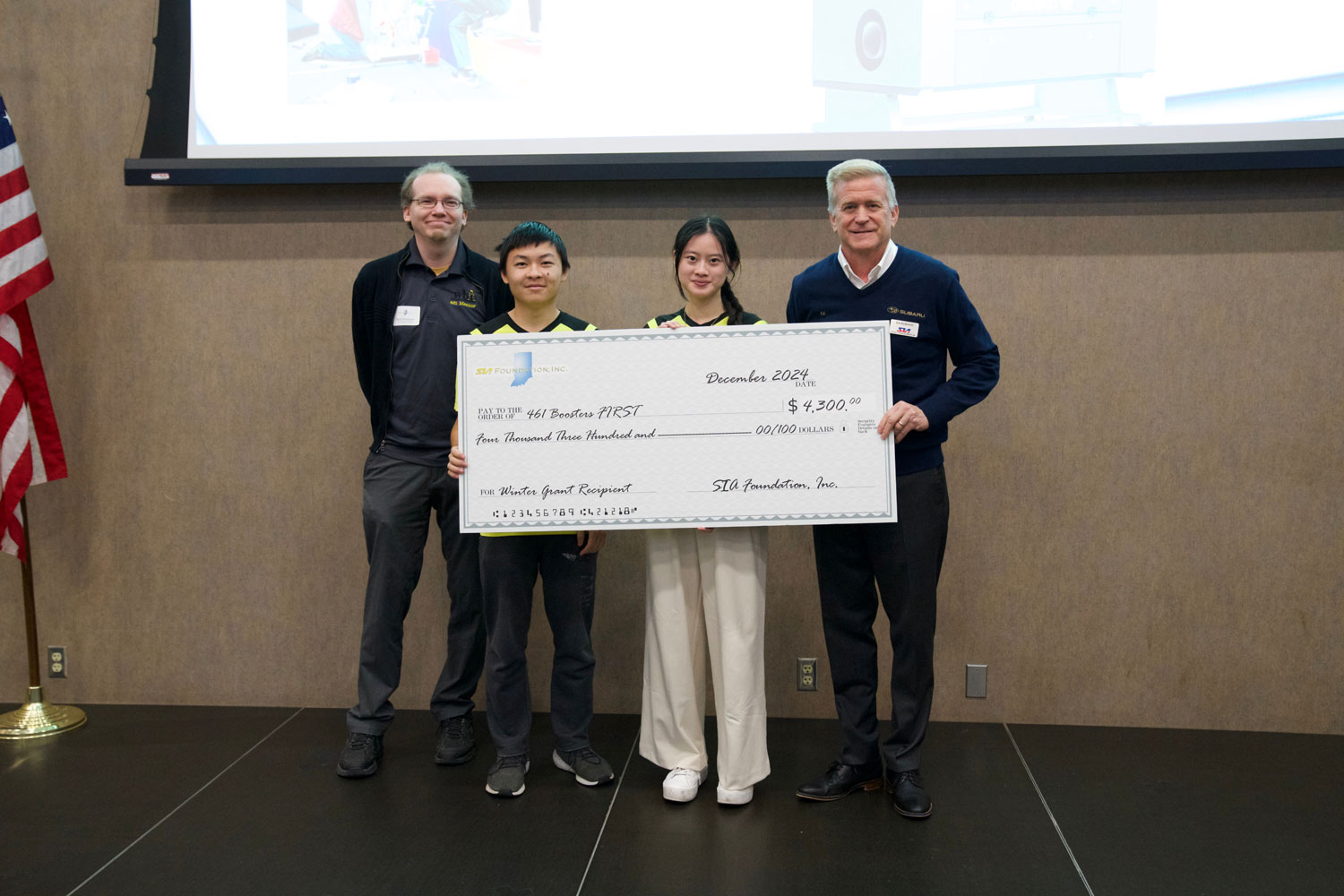 Four people stand together holding a large ceremonial check for $4,300. They're smiling, with a U.S. flag and a presentation screen in the background.