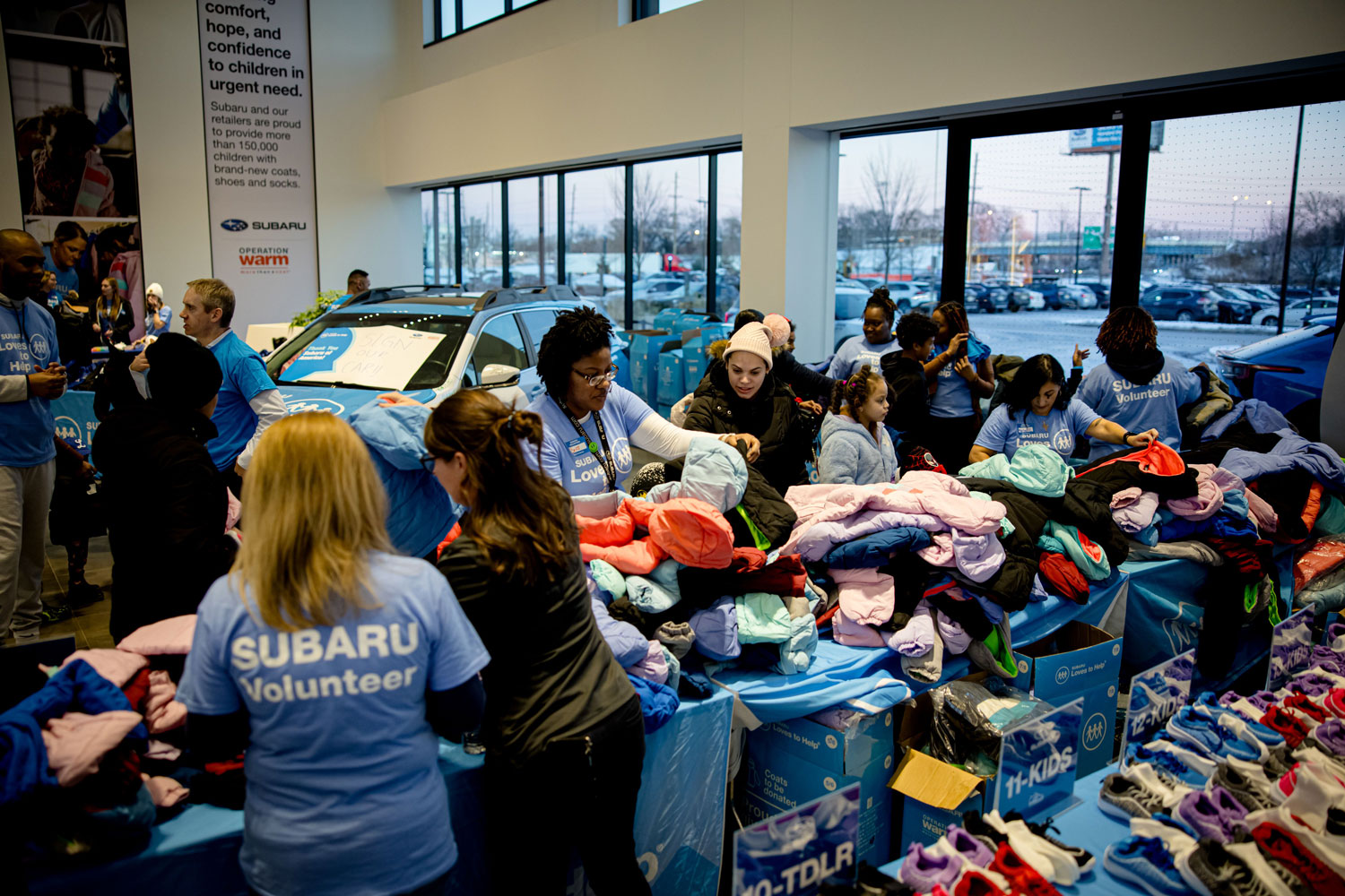 Volunteers in blue Subaru shirts distribute colorful winter coats and shoes to children inside a bright room.
