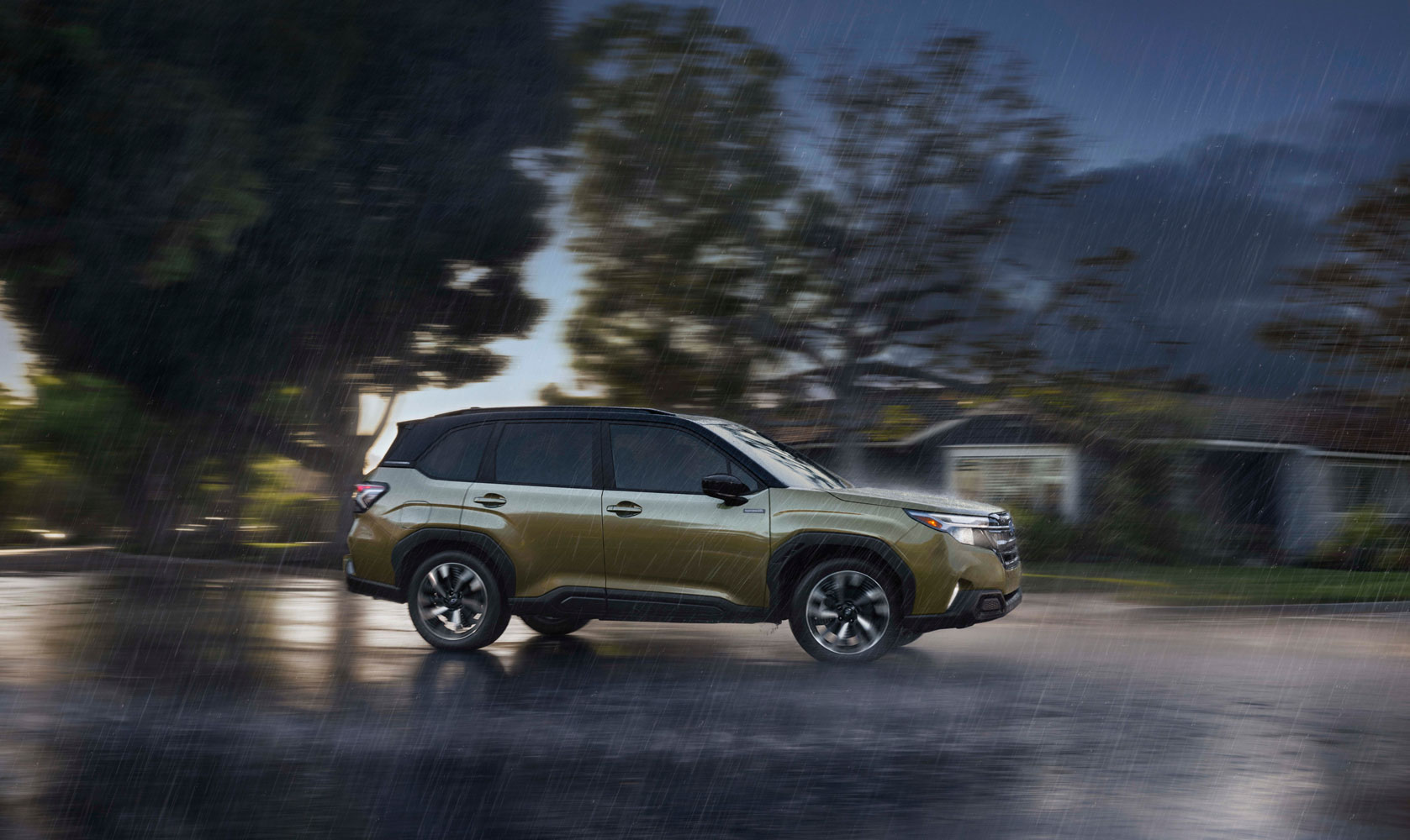 A metallic green Subaru SUV drives through heavy rain on a wet road. The background is blurred with trees and a house.