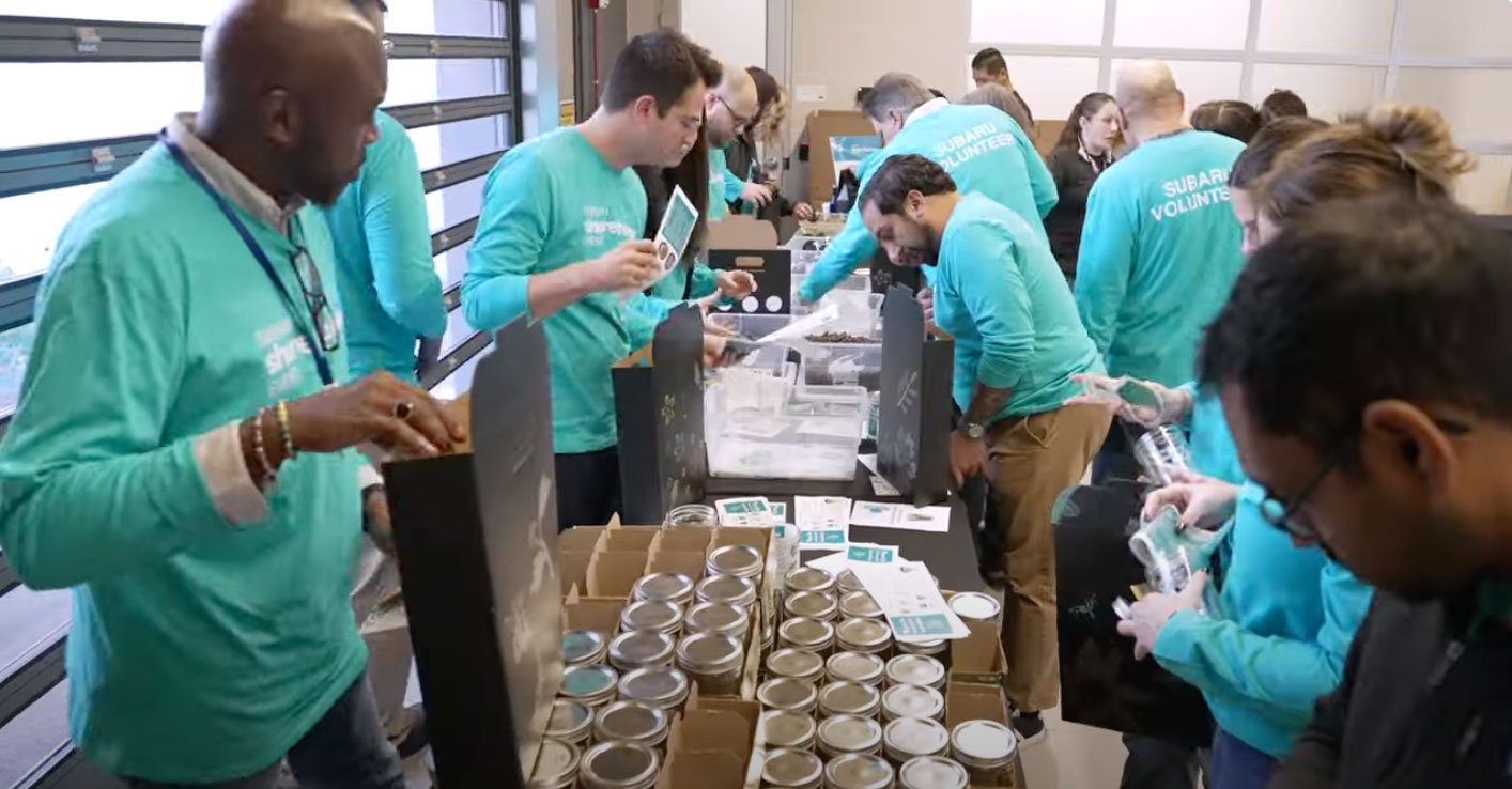 Volunteers in blue Subaru shirts pack items at a table filled with jars and supplies.