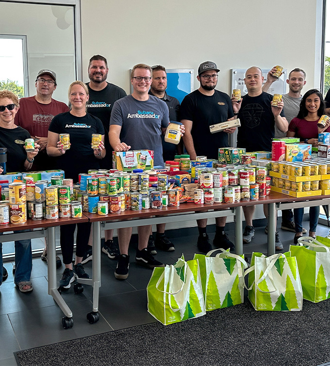 A group of smiling people holding cans stand behind a table filled with canned goods and food items. Reusable bags with green tree designs are in front.