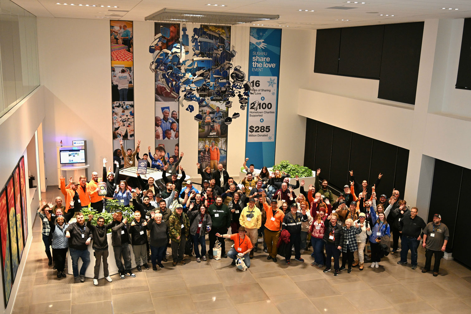 A large group of people stand in a brightly lit room, joyfully raising their hands. Behind them, banners display charity achievements and community images.