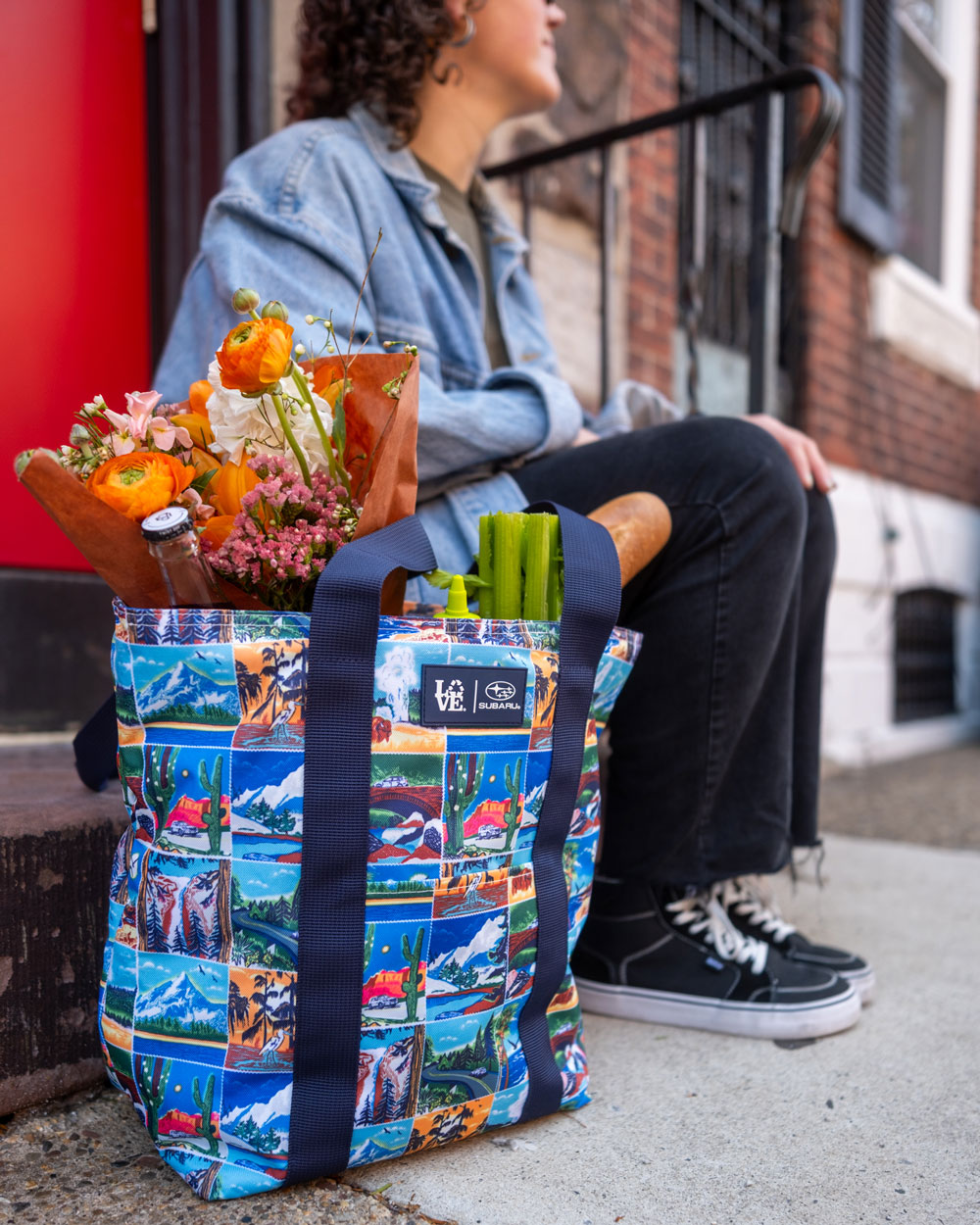 A vibrant shopping bag with National Park illustrations holds flowers, celery, and a baguette, sitting on a sidewalk next to a person in dark jeans and Vans.