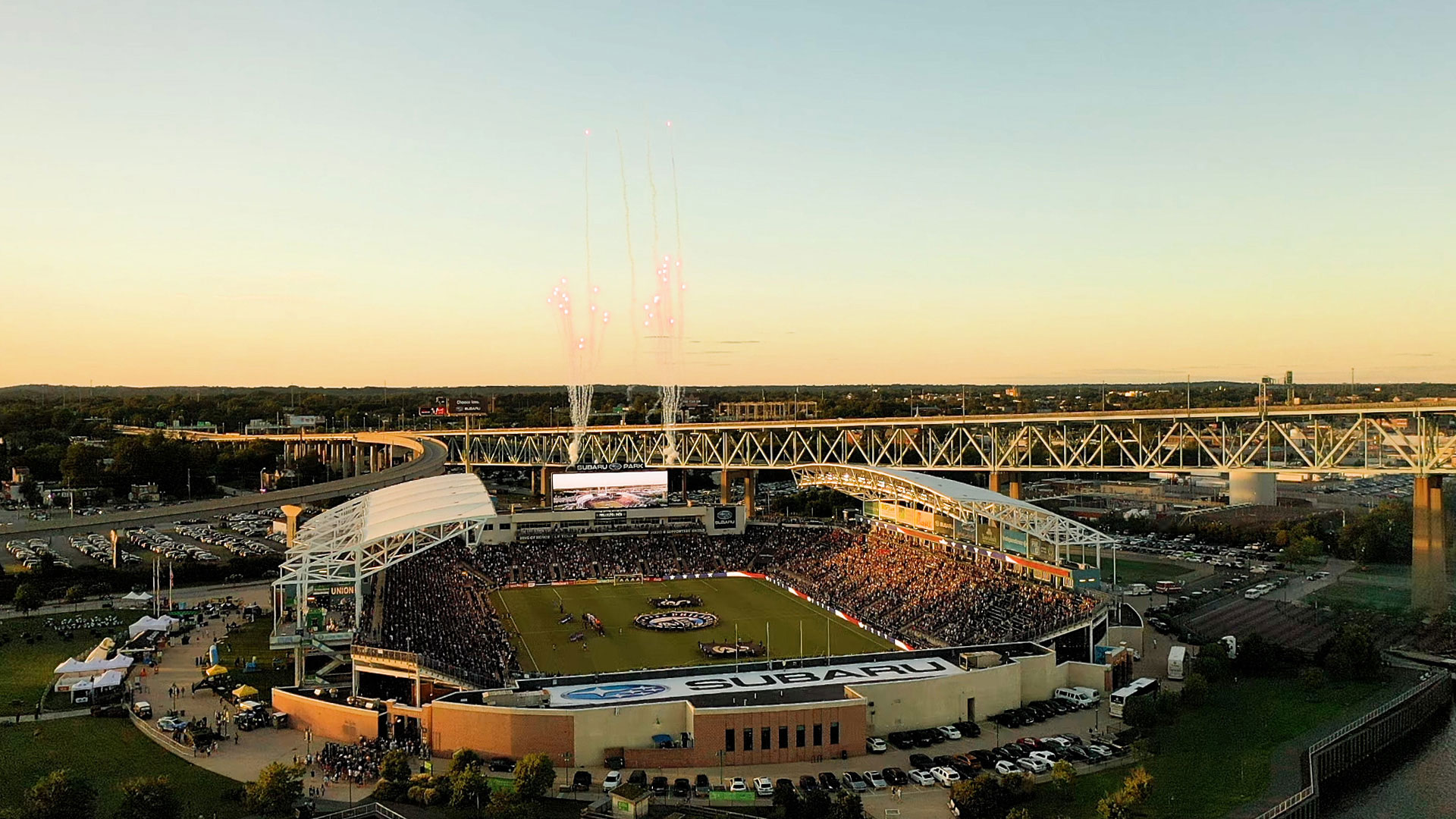 Aerial view of a packed Subaru Park stadium at sunset with a brightly lit scoreboard, surrounded by parked cars and a bridge in the background.