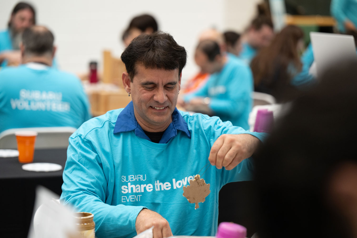Smiling man in a blue "Subaru Share the Love Event" shirt holds up a craft item. Background shows others in similar attire at a volunteer event.