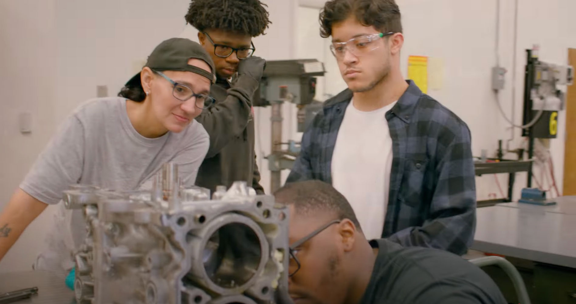 Four young adults in safety goggles focus intently on assembling a metal engine block in a workshop.
