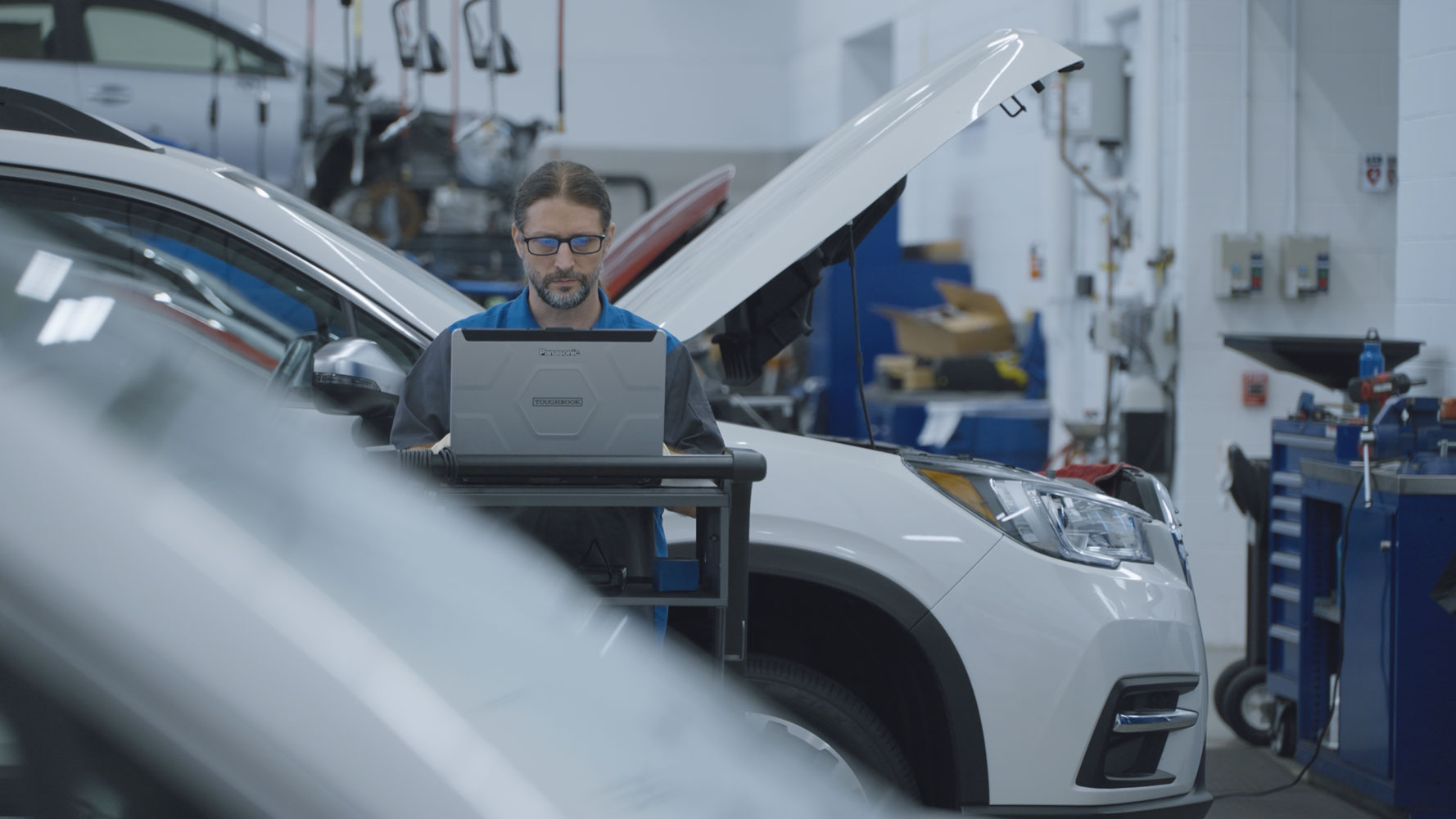 A mechanic with glasses works on a laptop in a car repair shop. A white SUV with an open hood is in the background.