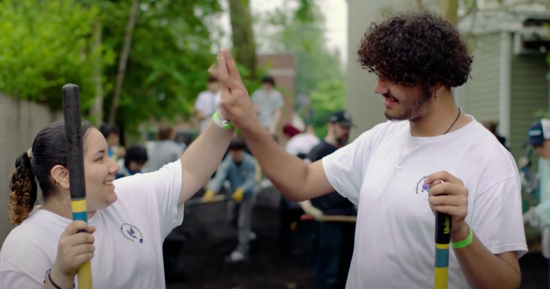 Two people in white shirts high-five outdoors, holding gardening tools, surrounded by a group working on a community project.