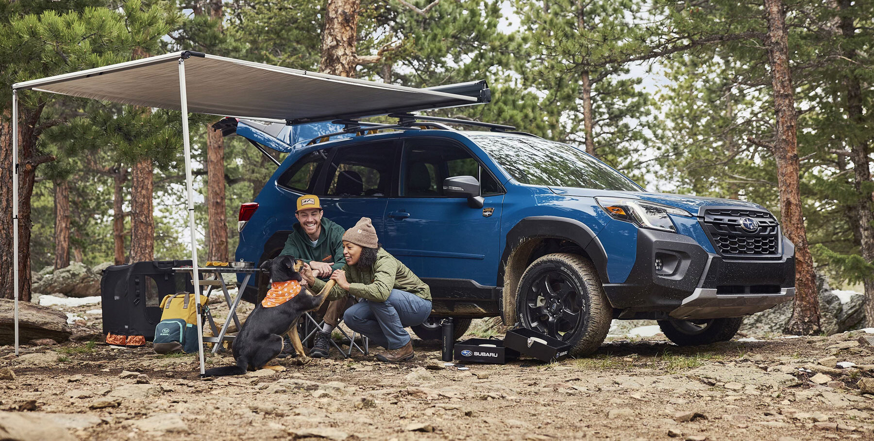 A couple smiling while playing with a dog in front of a blue Subaru SUV and camping gear in a forest setting.