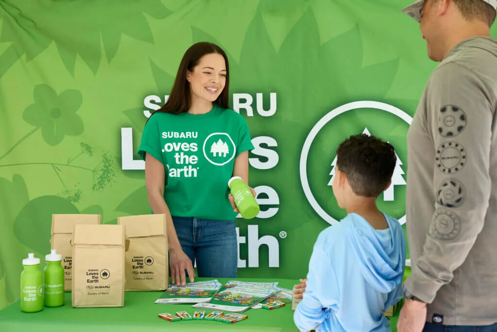 Three Subaru employees at a stewardship event table stacked with reusable paper bags and notebooks