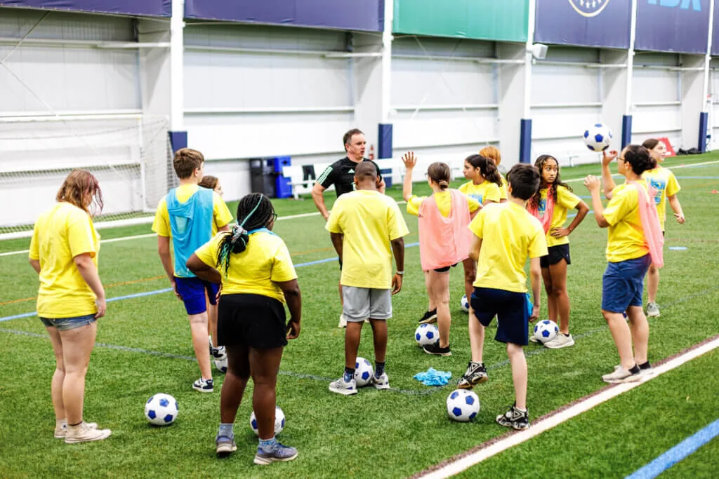 Youth soccer clinic at Subaru Park, with kids in yellow shirts gathered around a coach on the field