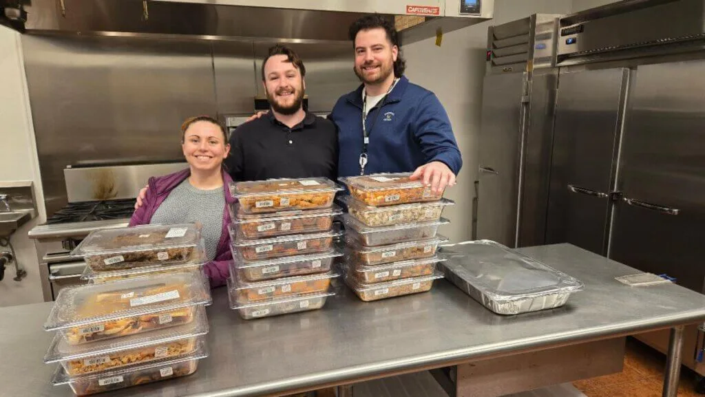 Three Subaru volunteers in a commercial kitchen with stacks of packaged meals prepared for the Share My Meals donation program