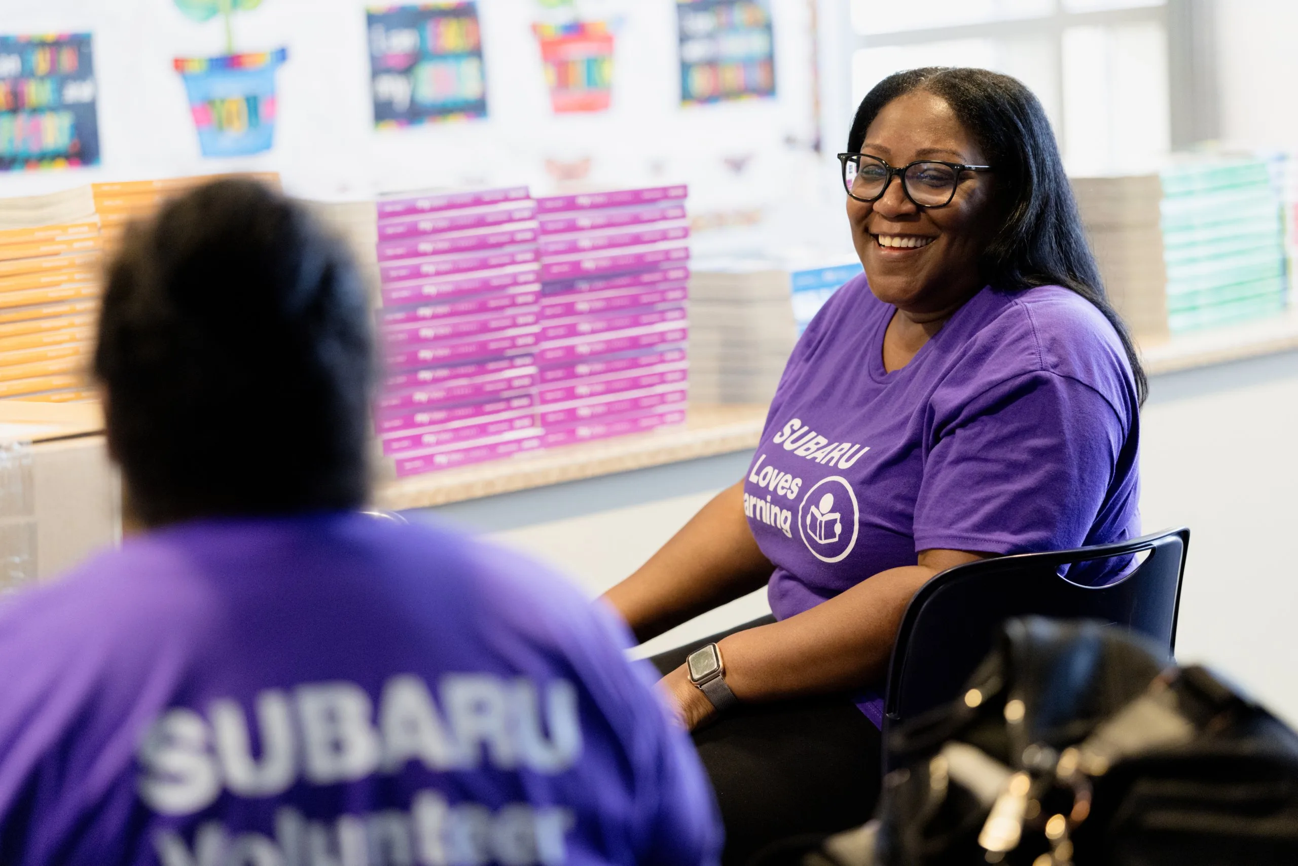 Subaru volunteers in purple Loves Learning shirts smiling together in a classroom stocked with books