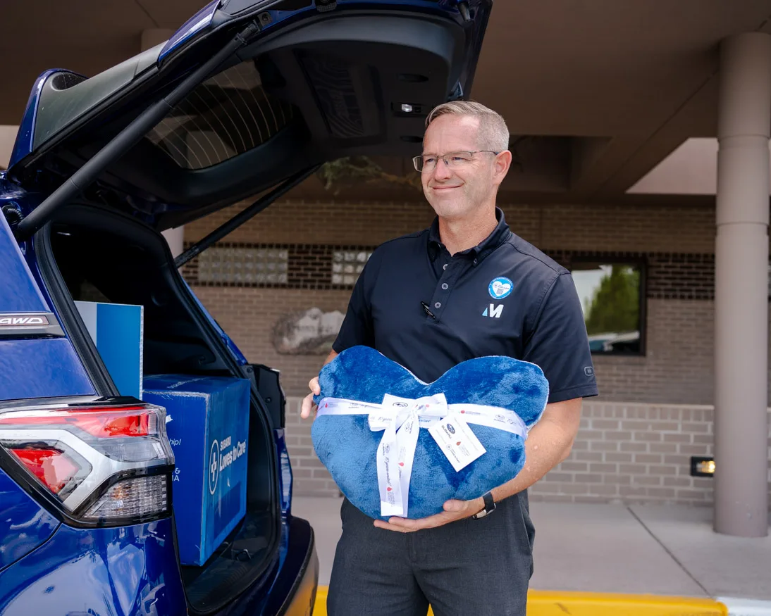 Subaru employee outside a hospital holding a heart-shaped pillow with messages of support, beside a Subaru Loves to Care vehicle