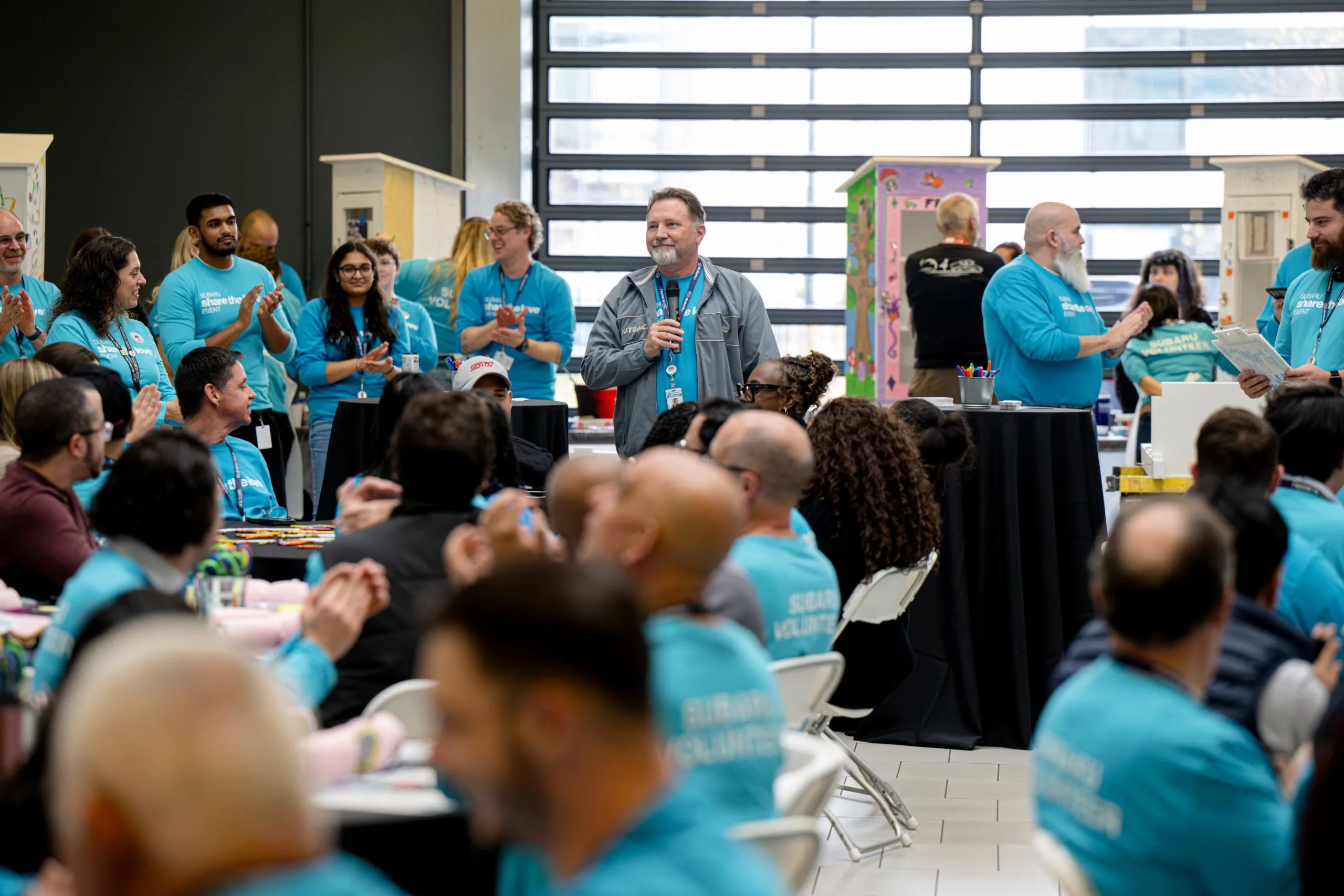 Subaru employee speaking into a microphone at a company volunteer event, surrounded by teammates in blue Subaru Volunteer shirts